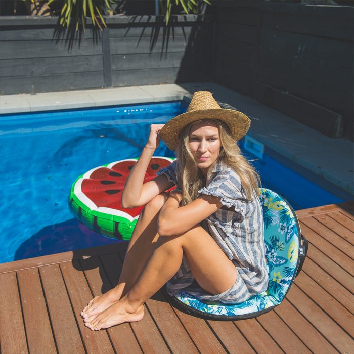 A woman sits poolside holding her sunhat, on a leaf print padded portable cushion recliner seat. The chair has an attractive tropical print in teal, blue, green yellow and white.