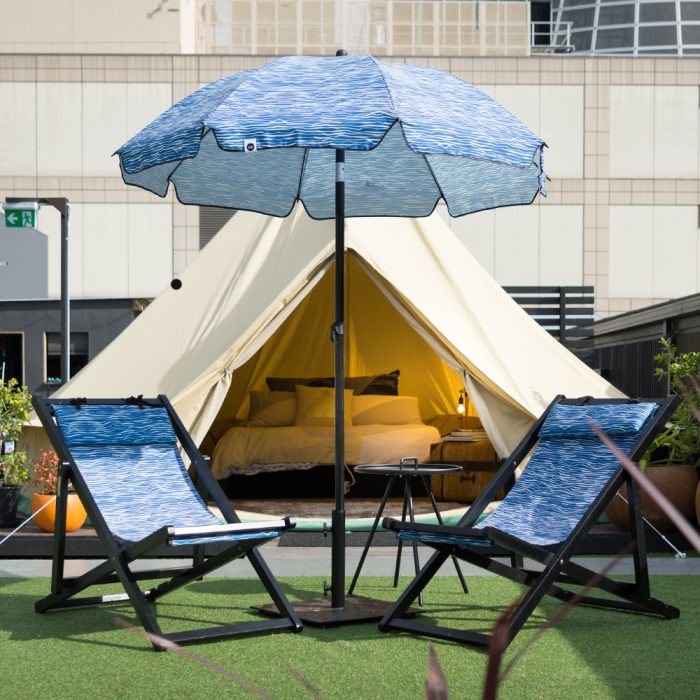 A blue wave print Wellen sun umbrella sits in front of a glamping tent with two matching deck chairs