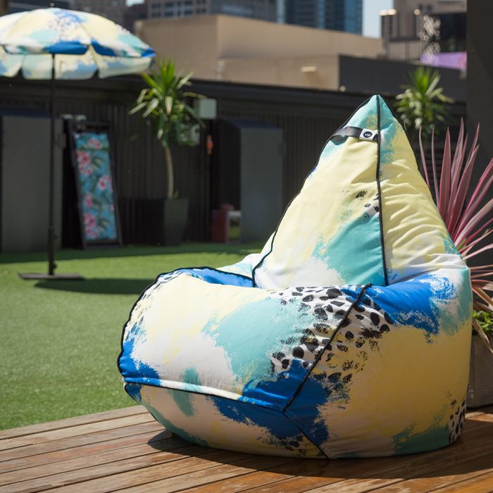 Tear drop shaped bean bag in bold abstract hand drawn designer tier fabric in green, blue, yellow and white with black splotches. Bean back is on a deck with a matching umbrella in the background