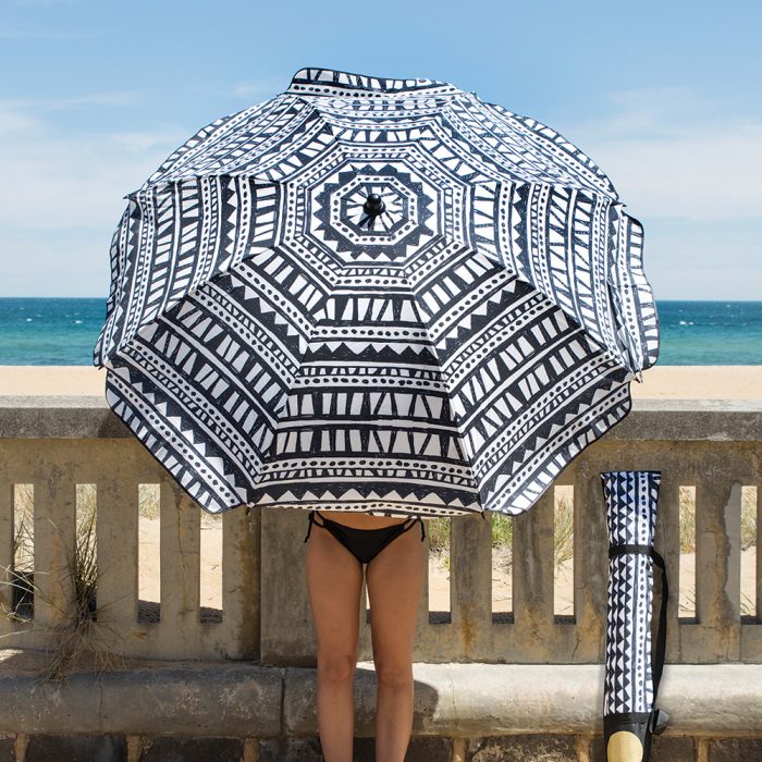 A woman holds the black and white geometric print bermuda sun umbrella leaning against a low concrete wall with a beach in the background