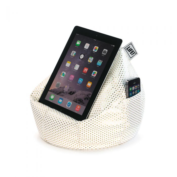 A tablet sits on a white iCrib tablet bean bag book rest with small metallic gold dot print fabric. A mobile phone is in the storage pocket.