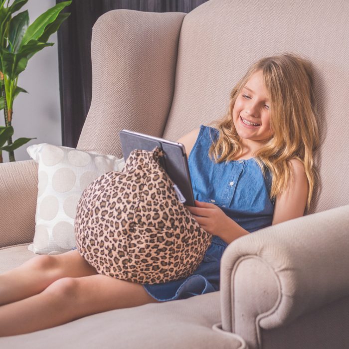 A teen sits on a couch reading from her iPad or tablet that sits in her lap nestled on a brown animal print iCrib bean bag.