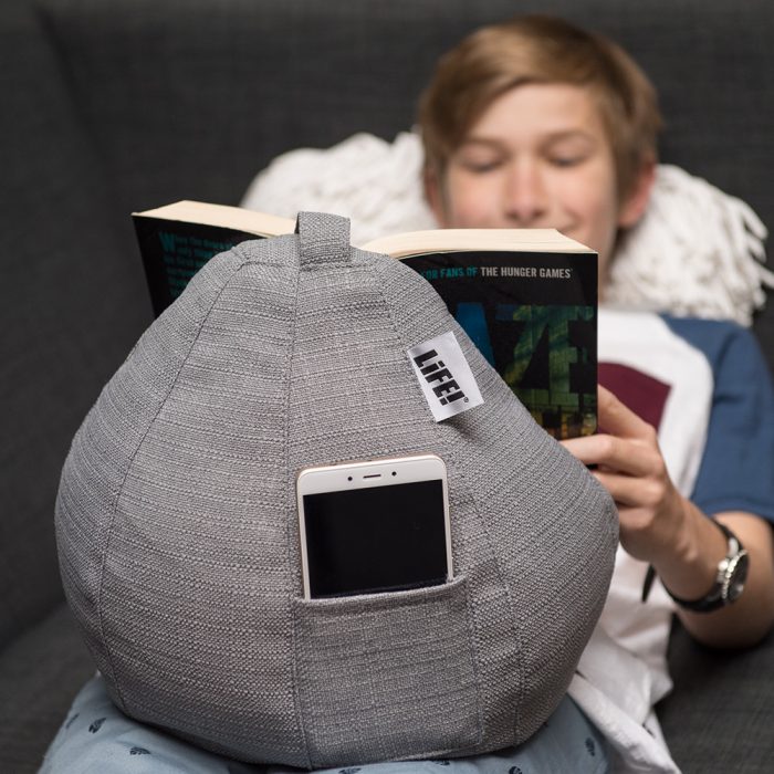 A teen reads a book resting on a grey linen look iCrib tablet holder. His phone sits in the storage pocket