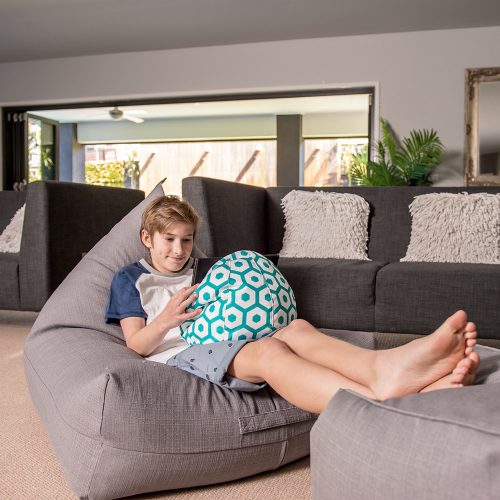 Teen sits on a grey linen look luna bean bag with his feet on a matching grey linen look ottaman watching his electronic device on a geo mint print iCrib