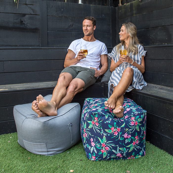 A couple relax drinking wine with their feet up on a grey linen look ottoman and blue tropical print ottoman