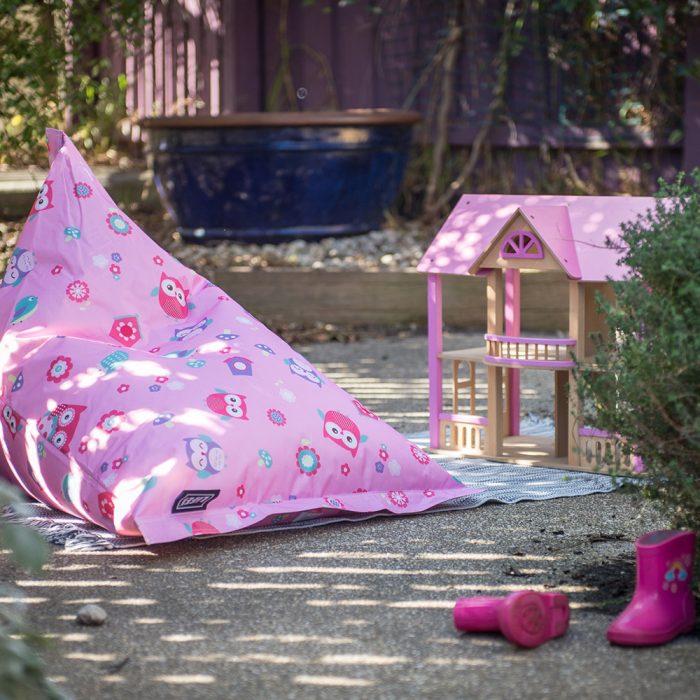 A sunny boy shaped kids bean bag sits in the garden outside next to a pink dollhouse and pink gumboots. The bean bag has a pink background with owls and flowers.