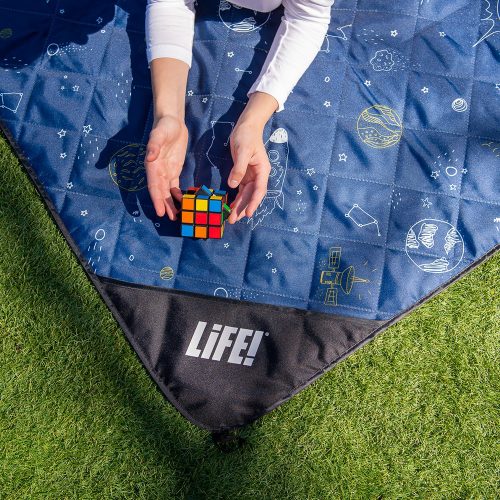 A teen plays with a rubics cube on a blue space buddy picnic rug. It has planets, stars and rockets printed in white