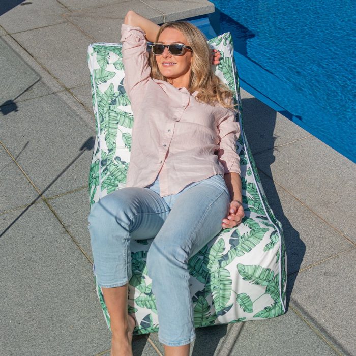 A woman reclines in the sunshine relaxed in a leaf tiki print coastal lounger bean bag by the pool
