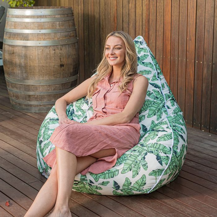 A woman sits on a super size tear drop bean bag with leaf print on her patio. The contrast white trim is visible.